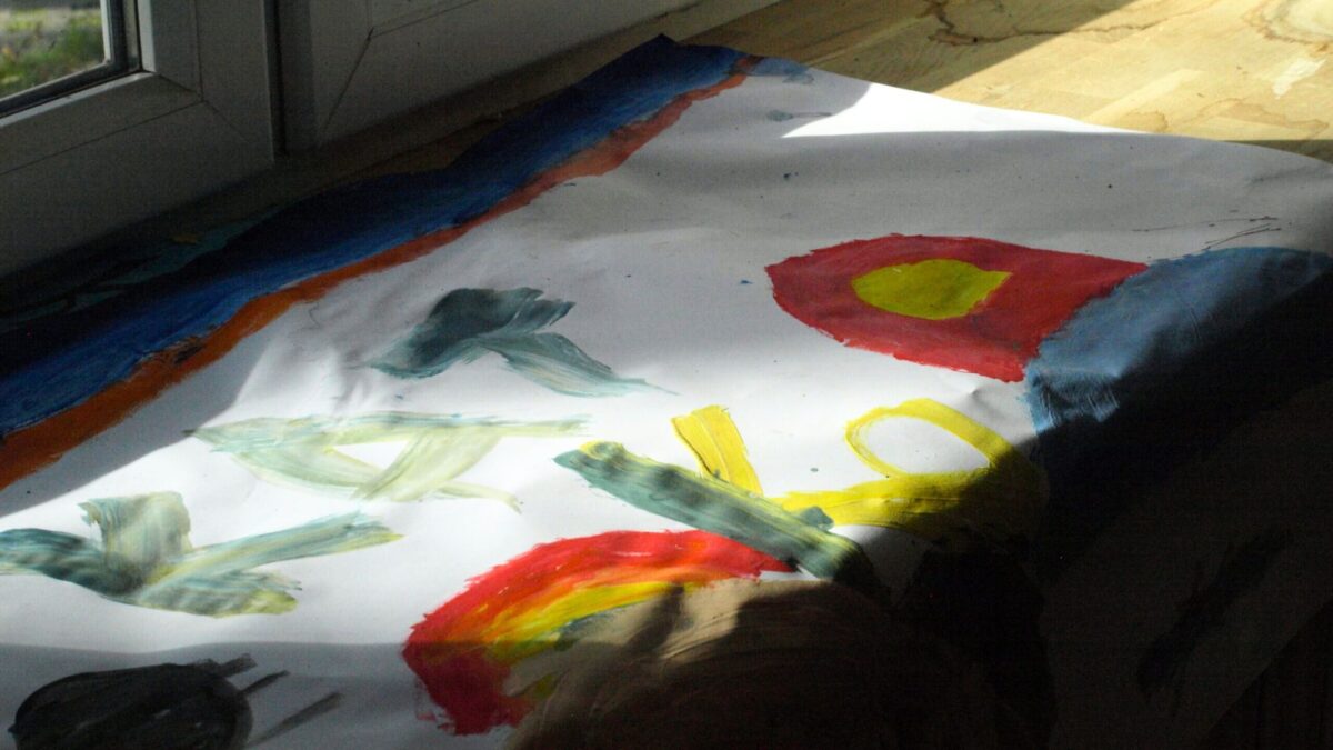 Biała kartka z kolorowym rysunkiem jednego z dzieci ze świetlicy w Dębaku. Rysunek został wykonany farbami. Teraz leży na parapecie, susząc się. Częściowo oświetla go światło z okna, a częściowo znajduje się w cieniu.